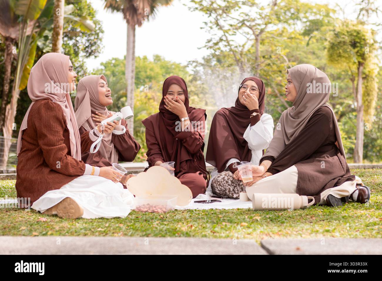 Un gruppo di giovani donne musulmane in hijab si siedono insieme all'aperto, condividendo risate e abbracciando l'amicizia. La scena cattura l'essenza di un moderno, Foto Stock