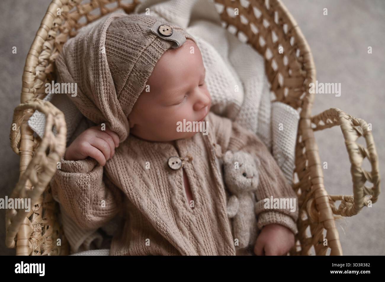 Un bambino appena nato tranquillo, vestito con un intimo abito a maglia, si trova comodamente in un cestino in tessuto. Il bambino tiene in mano un piccolo peluche, creando un sorriso Foto Stock
