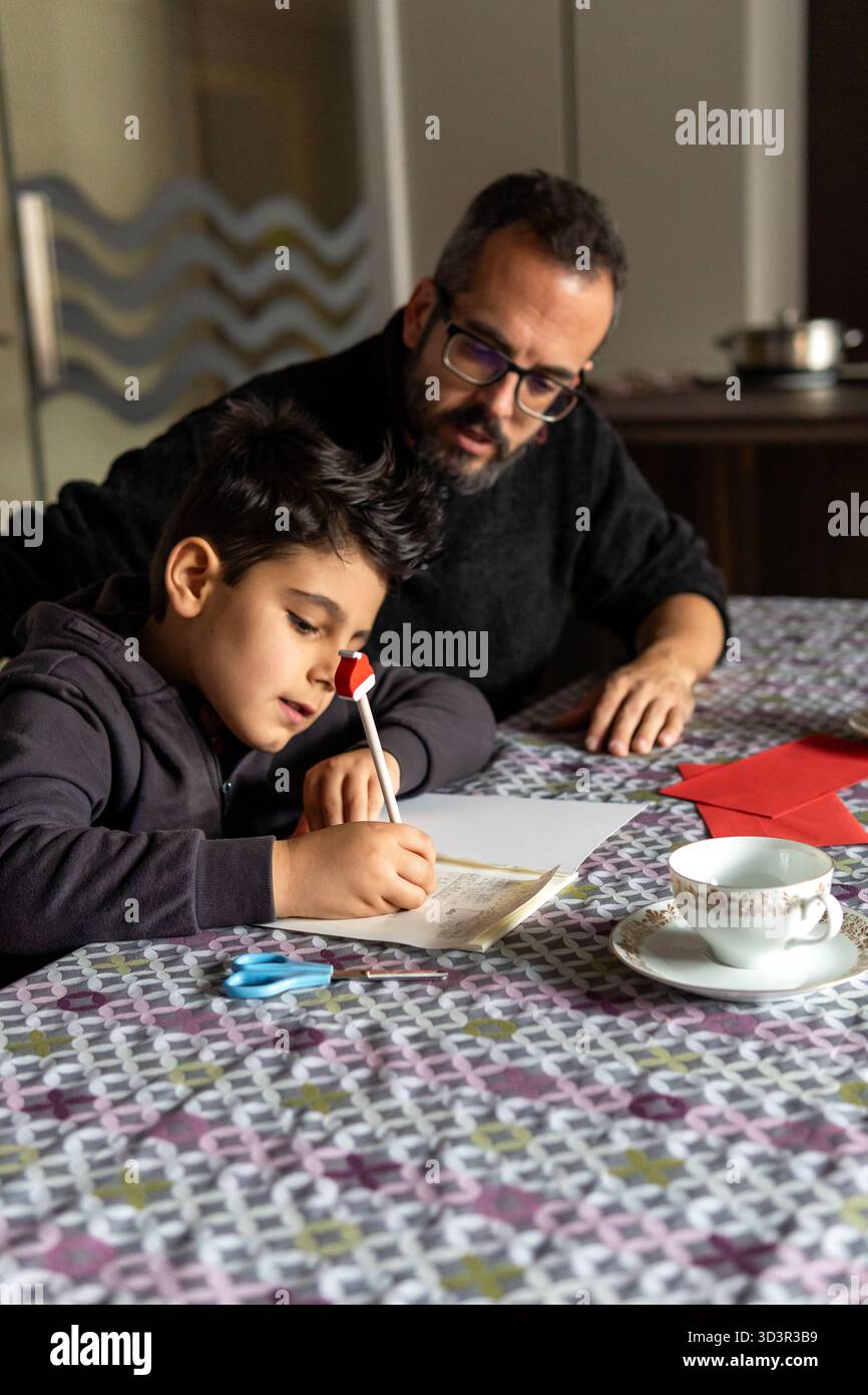 Padre e bambino siedono a un tavolo colorato, concentrandosi sulla scrittura di una lettera di cuore a Babbo Natale e ai tre saggi, circondati dal festoso dicembre Foto Stock