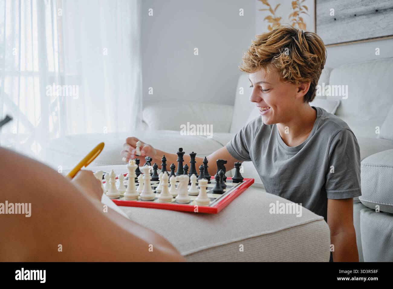 Adolescente sorridente impegnato in una partita amichevole di scacchi a casa, mostrando concentrazione e divertimento. La luce naturale mette in risalto l'ambiente accogliente e confortevole Foto Stock