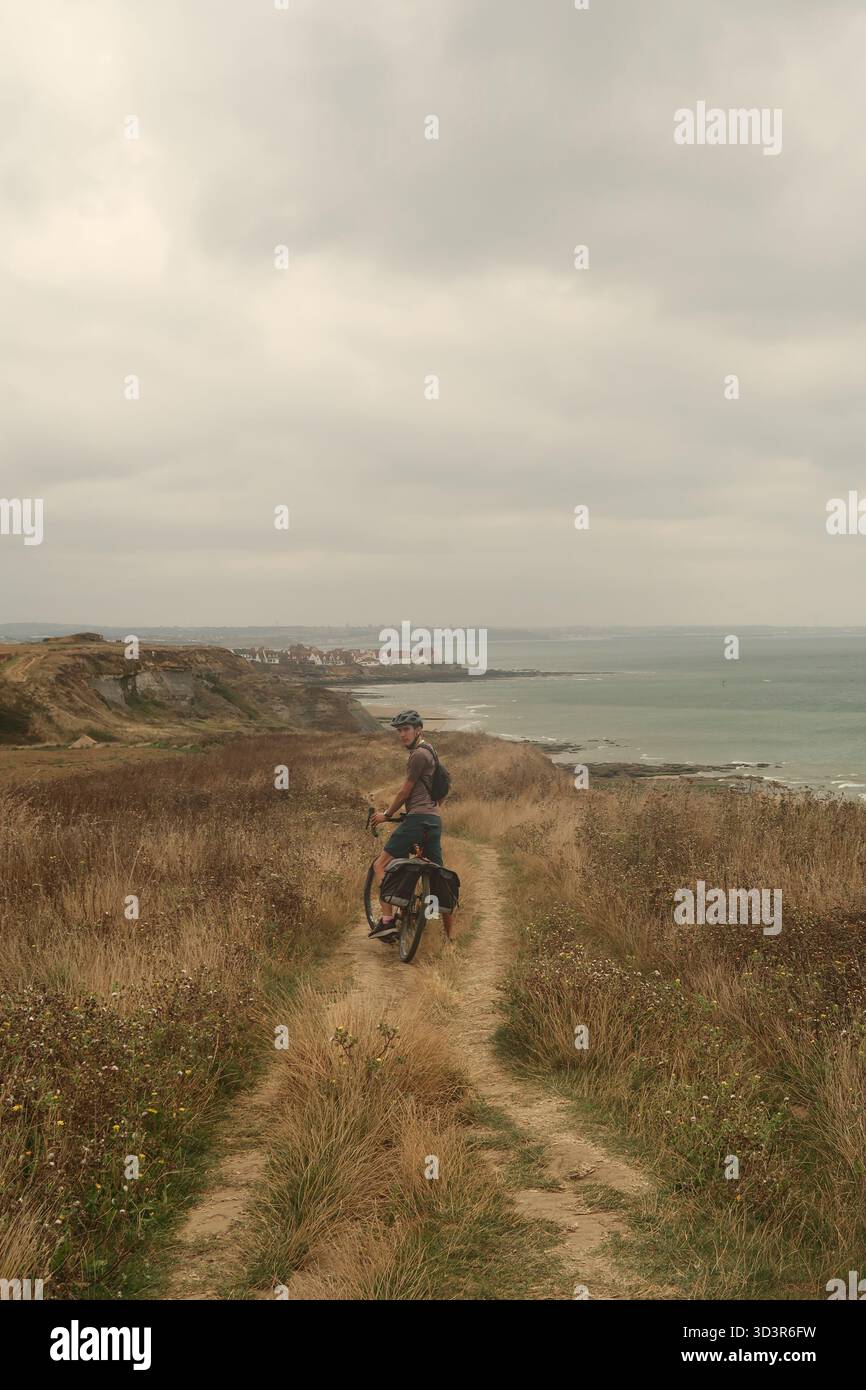 Un ciclista solitario percorre un sentiero accidentato lungo la splendida costa settentrionale della Francia. Il paesaggio sereno, caratterizzato dai suoi campi dorati ed expa Foto Stock