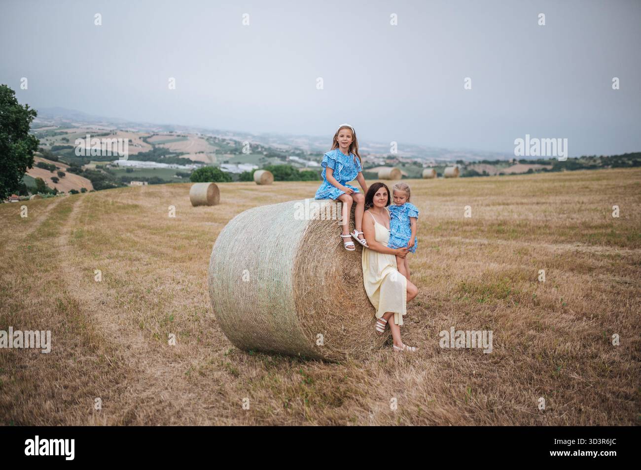 Un momento sereno in un campo di campagna italiano vede una madre e le sue figlie sedute su una balla di fieno. Le colline ondulate e i cieli limpidi creano una pict Foto Stock