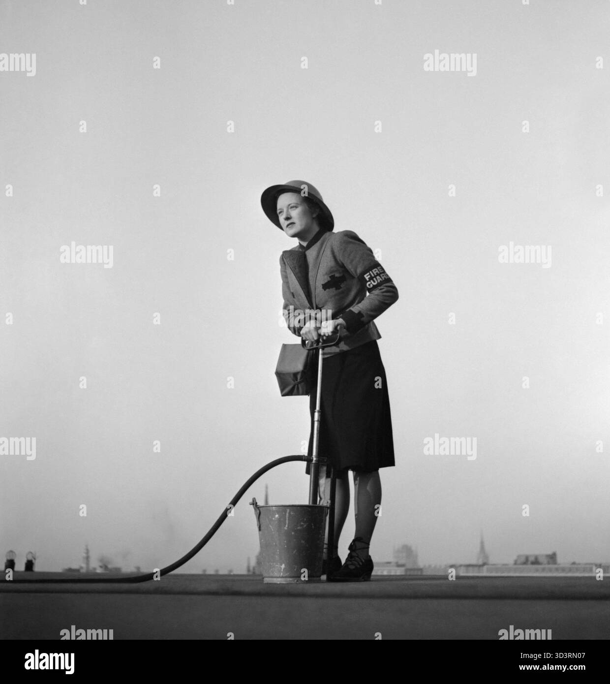 Londra, 1941. Una guardia del fuoco femminile sta su un tetto usando una pompa a staffa durante il Blitz. Indossando un casco d'acciaio, la fascia da braccio della Guardia antincendio e una maschera antigas, osserva le bombe incendiarie mentre lo skyline della città si trova dietro di lei. Probabilmente fotografato nell'ottobre 1941. Foto Stock