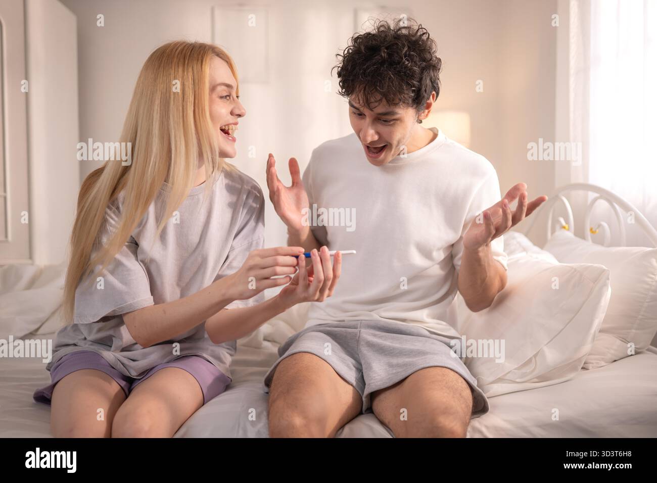 Coppia felice seduta sul letto e sorridente di eccitazione guardando un test di gravidanza positivo || modello rilasciato Foto Stock