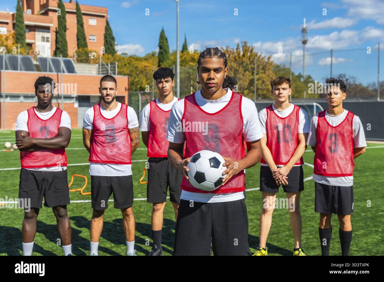 Giovani provenienti da diversi background che formano una squadra di calcio, in piedi su un campo di erba sintetica verde con un giocatore che tiene la palla, allenandosi per loro Foto Stock