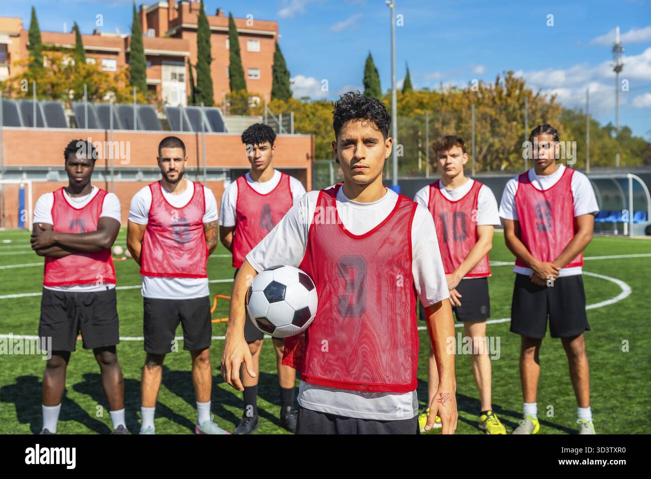 Gruppo eterogeneo di giocatori di calcio maschili in pettorali rossi in piedi su un campo di allenamento, tenendo in mano una palla e pronti per la pratica o per una partita competitiva con concentrazione Foto Stock