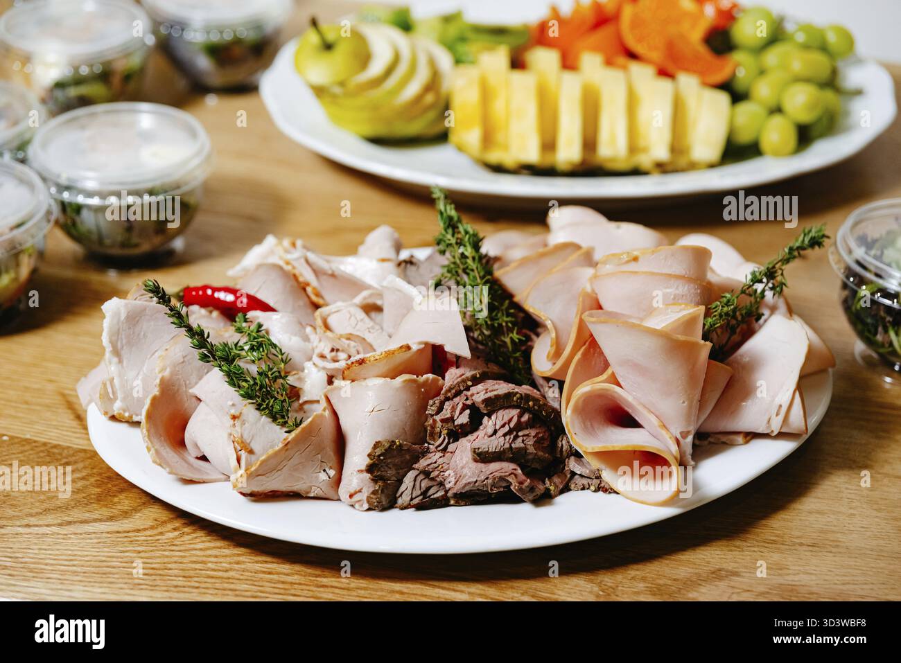 Cibo, Una varietà di affettati freddi serviti su un piatto, accompagnati da erbe e peperoncino, vassoio di frutta fresca sullo sfondo Foto Stock