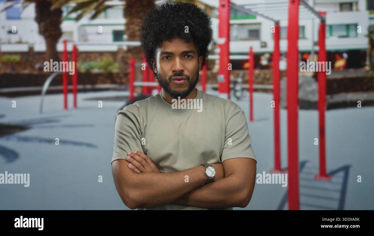 L'uomo con le braccia incrociate si trova in una palestra all'aperto circondata da attrezzature da palestra rosse, adagiata su uno sfondo urbano moderno con palme. Foto Stock
