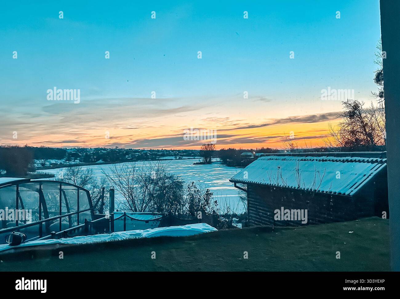 Splendida scena invernale che mostra un lago ghiacciato circondato da alberi e un rustico capannone in legno, con uno splendido tramonto che illumina il cielo in calde tonalità Foto Stock