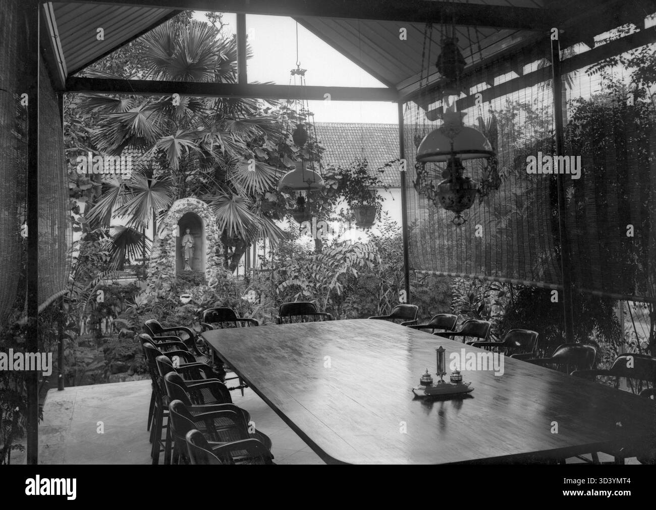 Storia coloniale in Indonesia (ex Indie Orientali olandesi) tra il 1895 e il 1910: Un grande tavolo da pranzo sotto un tetto, con vista su un giardino e una statua di un santo maschio o Gesù in una nicchia decorata con conchiglie, forse in una scuola missionaria. Foto Stock