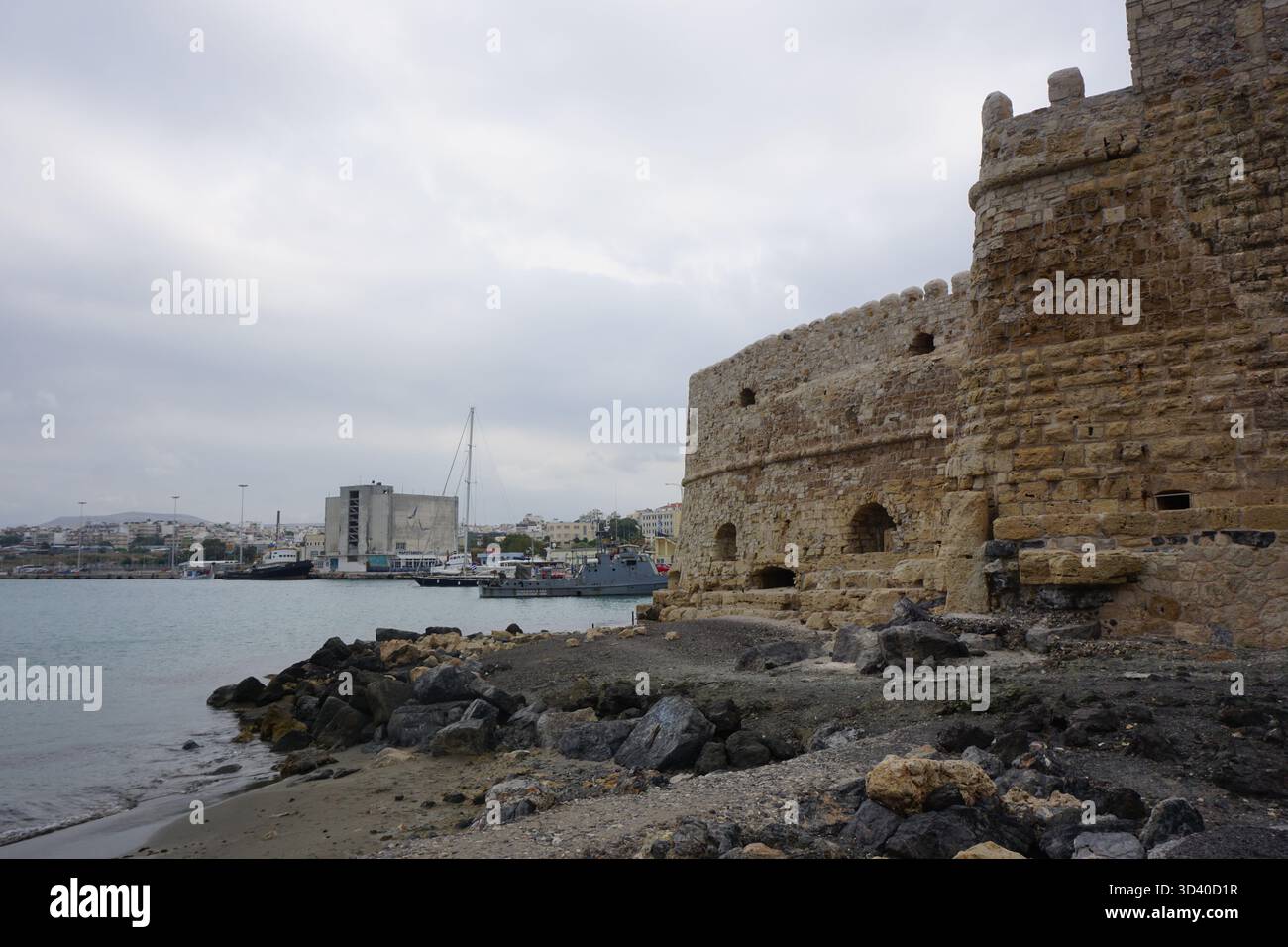 La Fortezza di Koules incontra la spiaggia rocciosa, con una motovedetta della Guardia Costiera greca e moderni edifici portuali visibili sullo sfondo, epoche contrastanti Foto Stock