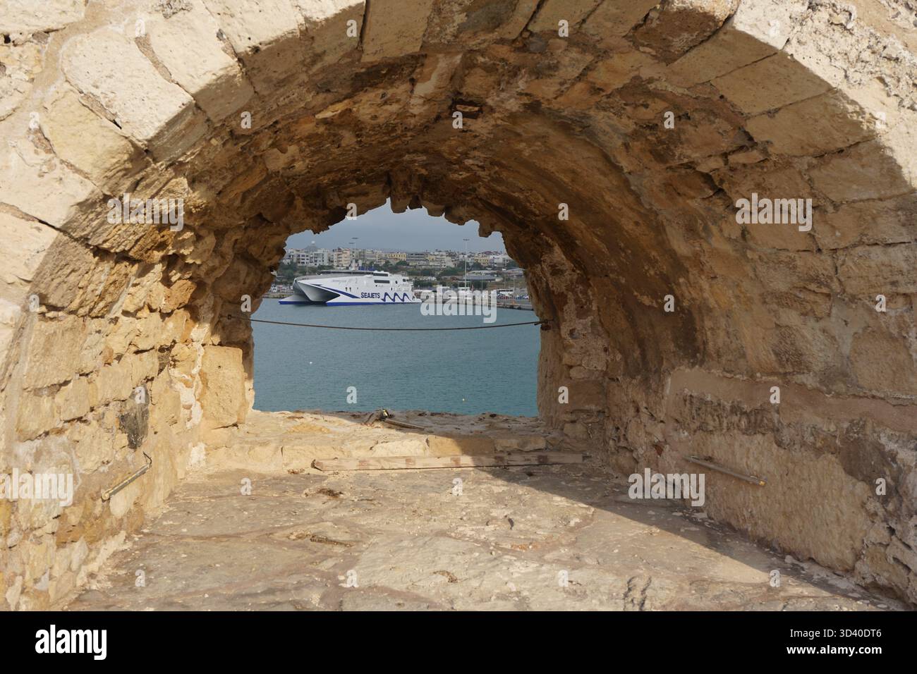 Un moderno catamarano passeggeri ad alta velocità attraversa il mare, incorniciato perfettamente dall'arco di pietra intemprato della storica fortezza di Koules a Heraklion Foto Stock
