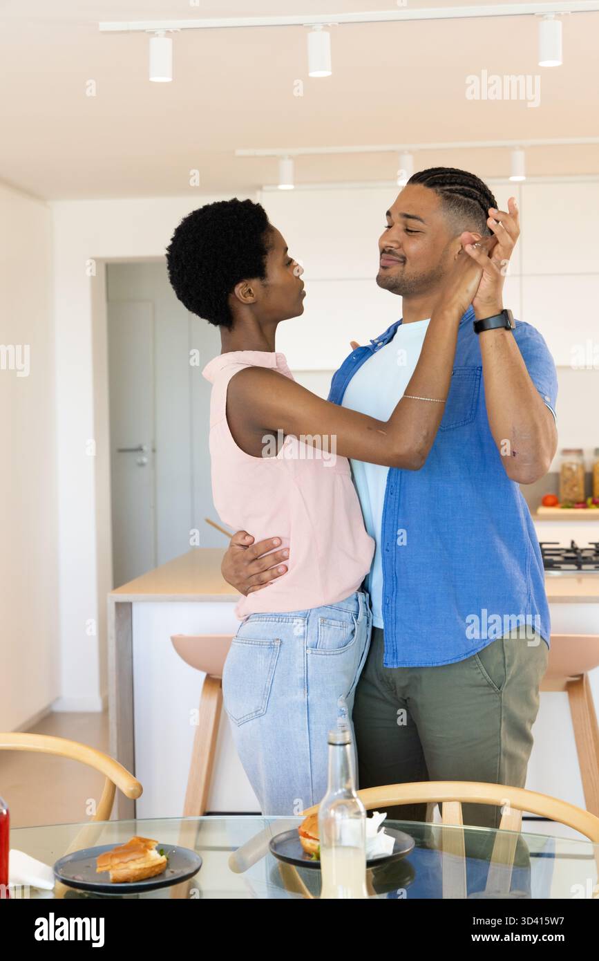 Coppia romantica e variegata che si trova vicino all'isola della cucina e a un tavolo con piano in vetro, con panini Foto Stock