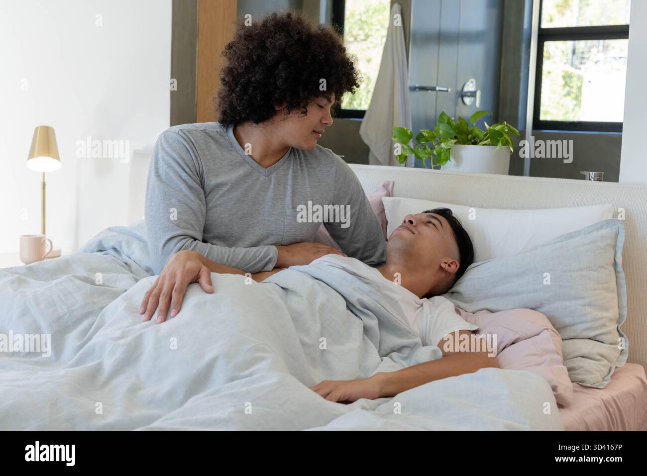 Appetitose coppie di uomini con biancheria da letto leggera nella moderna camera da letto, con lampada dorata e tazza rosa Foto Stock