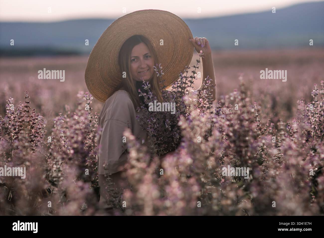 Campo dei fiori del cappello donna: Ritratto estivo, donna felice che indossa un cappello di paglia tra fiori viola in fiore in un campo, che evoca serenità. Foto Stock