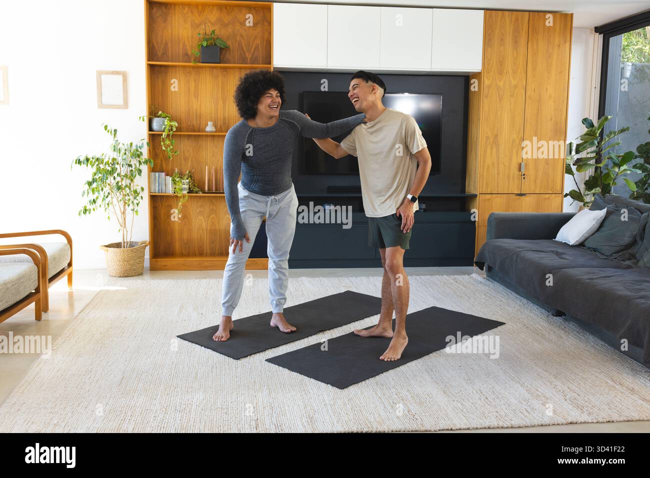 Amici maschi che ridono in piedi su tappetini neri da yoga nel moderno soggiorno, con scaffalature in legno Foto Stock
