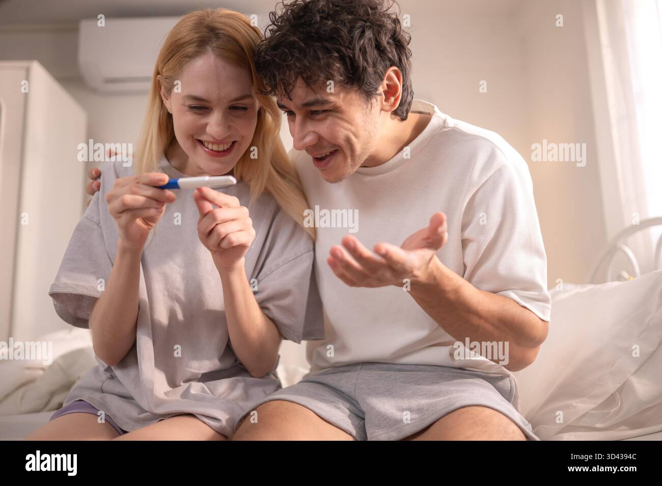 Coppia felice seduta sul letto e sorridente di eccitazione guardando un test di gravidanza positivo || modello rilasciato Foto Stock