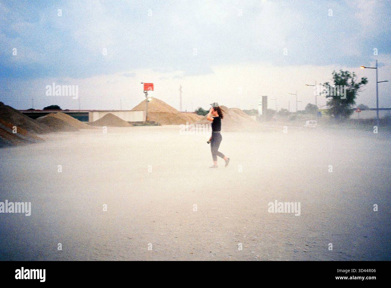 Ragazza che cammina in un cantiere di materiali da costruzione, soffia di vento e polvere mentre si protegge, indossa abiti neri, dinamica scena analogica. Foto Stock