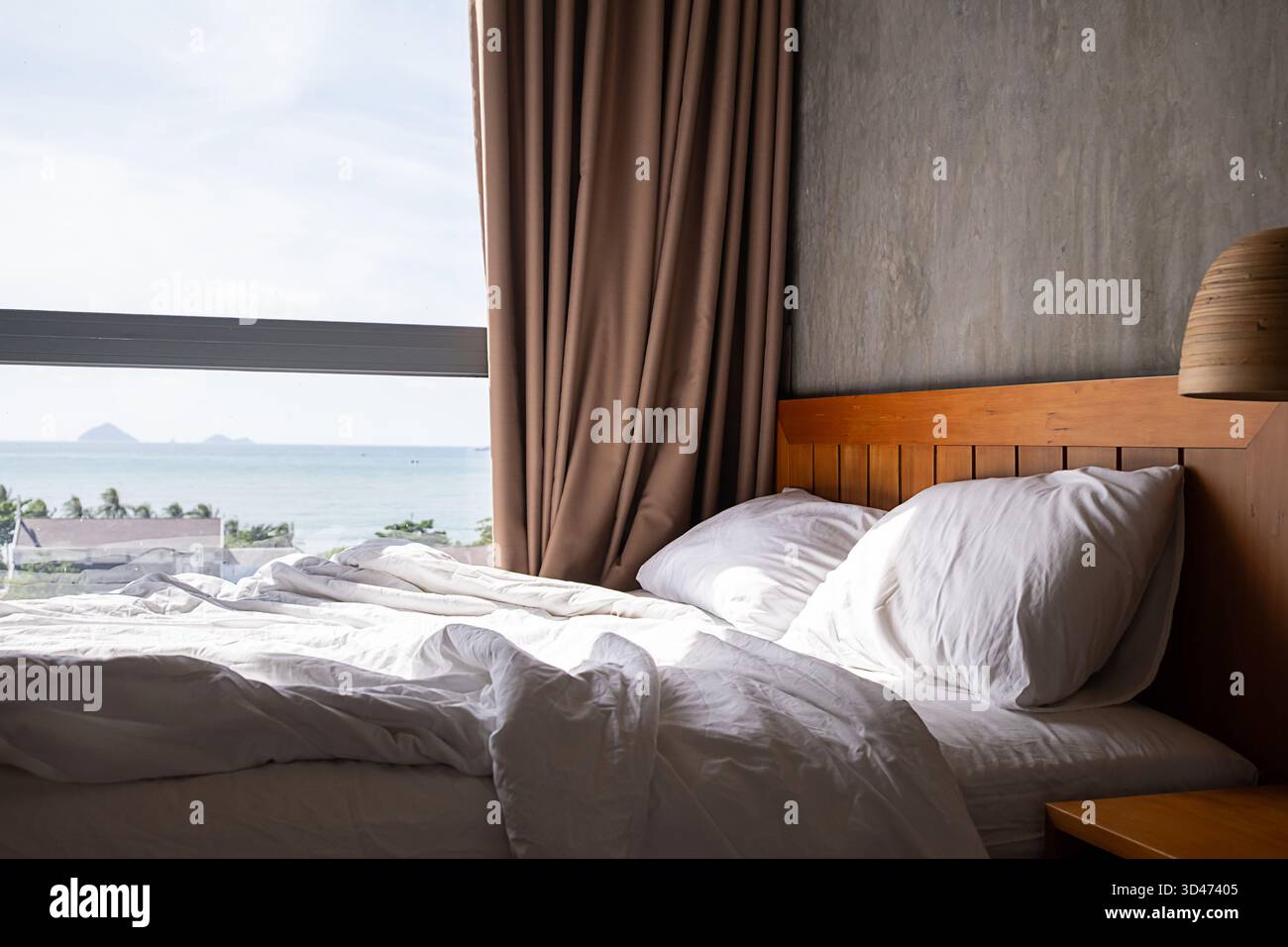 Comodo letto con lenzuola bianche e cuscino in camera soleggiata vicino a una grande finestra affacciata sul mare, perfetta camera da letto tranquilla per il relax. Foto Stock