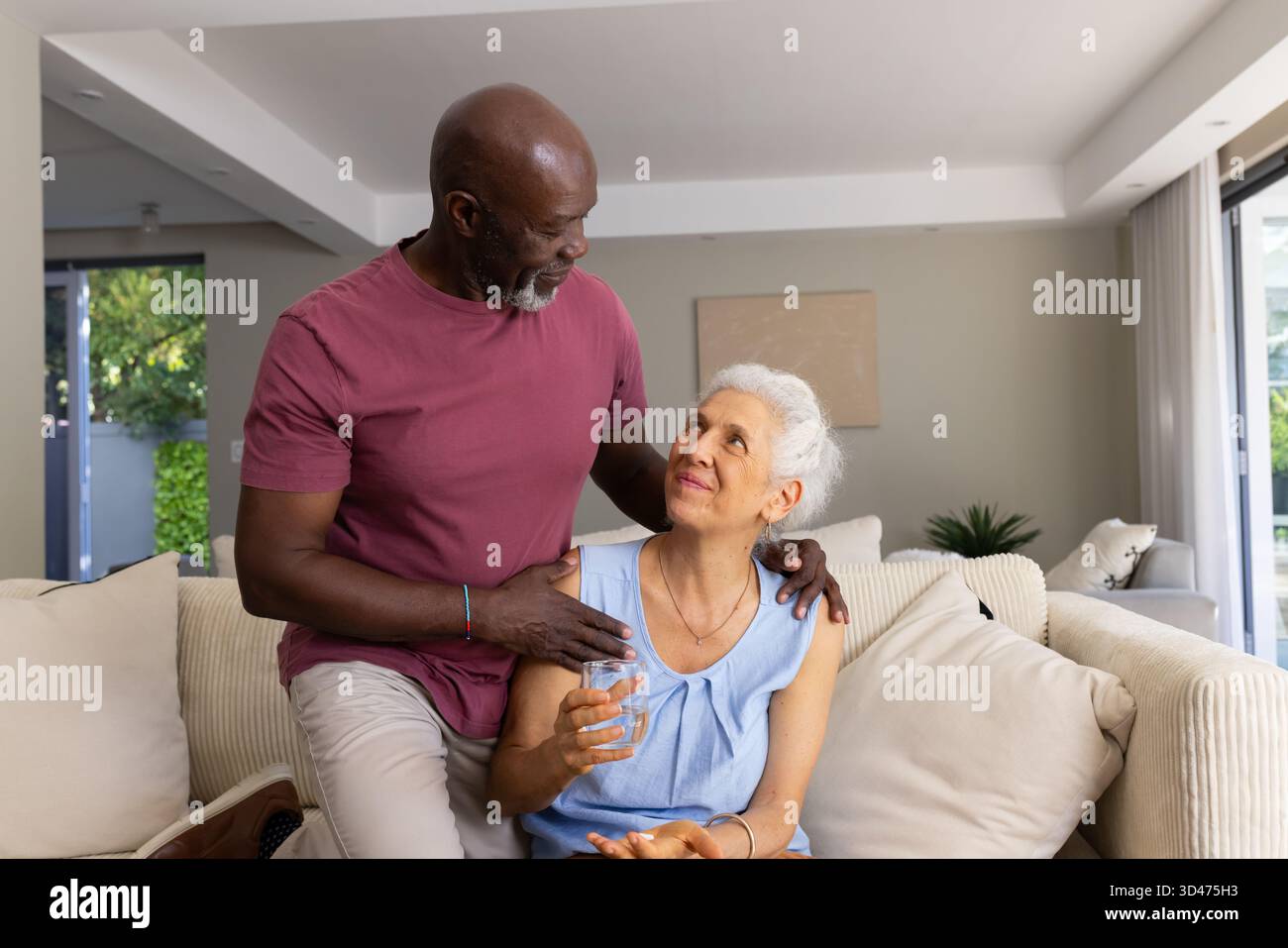 Senior uomo e donna afroamericana che condividono momenti sul divano di casa, con un bicchiere d'acqua Foto Stock