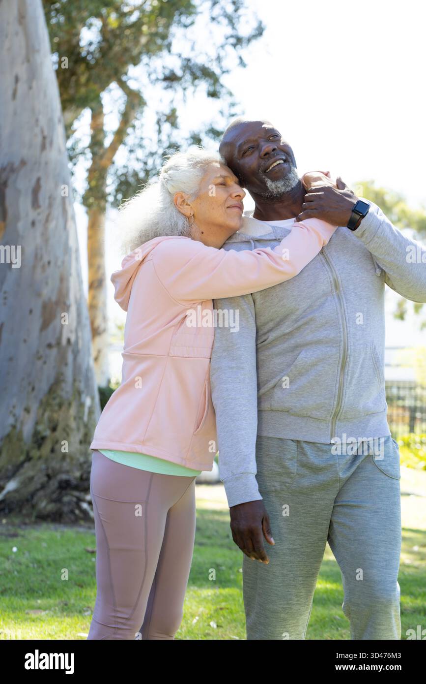 Diverse coppie senior che si abbracciano sotto un grande albero nel parco urbano, con smartwatch nero Foto Stock