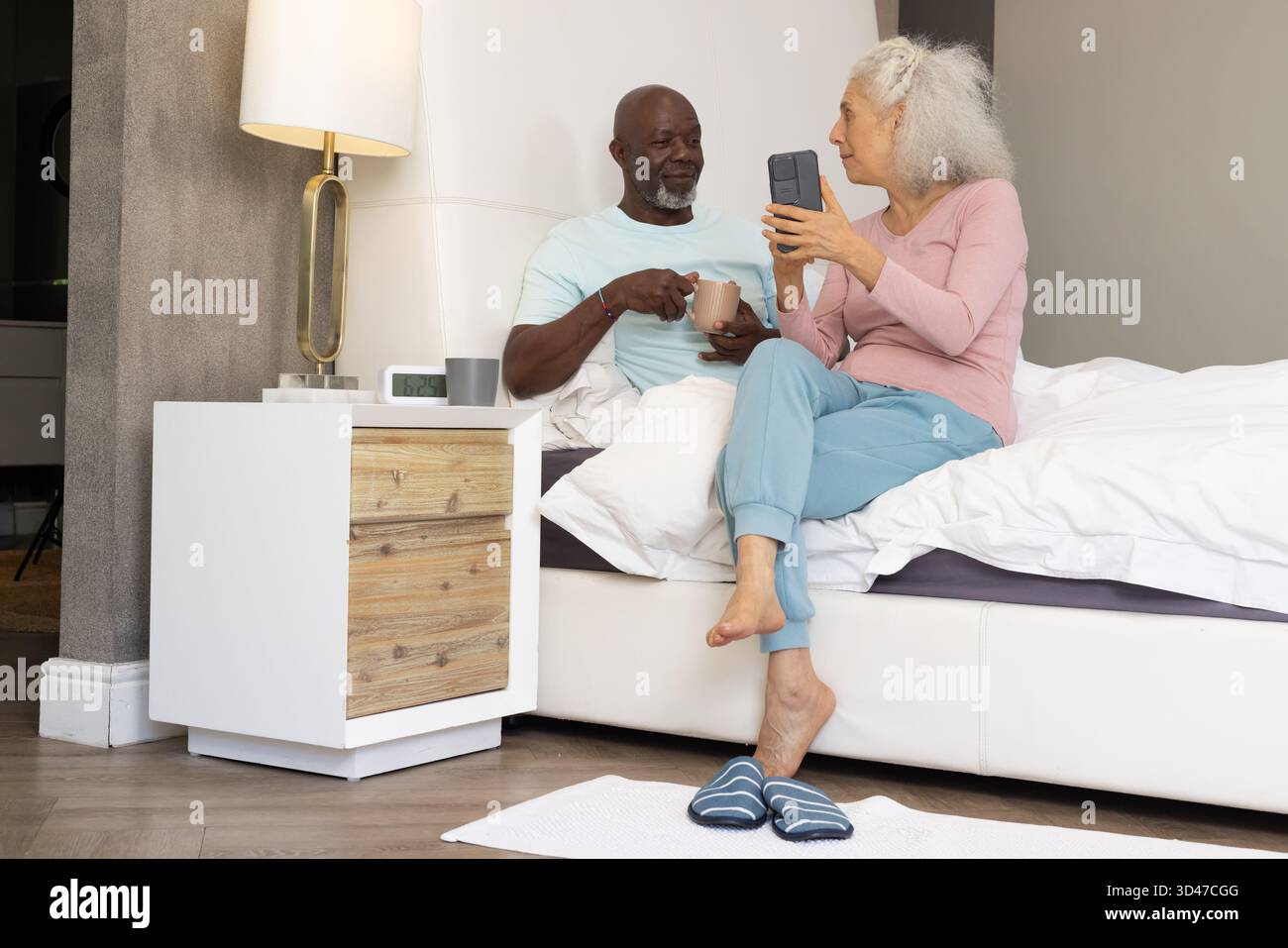 Sorseggiando un caffè, una coppia senior molto varia, seduta sul letto di casa, con smartphone e tazza da caffè Foto Stock