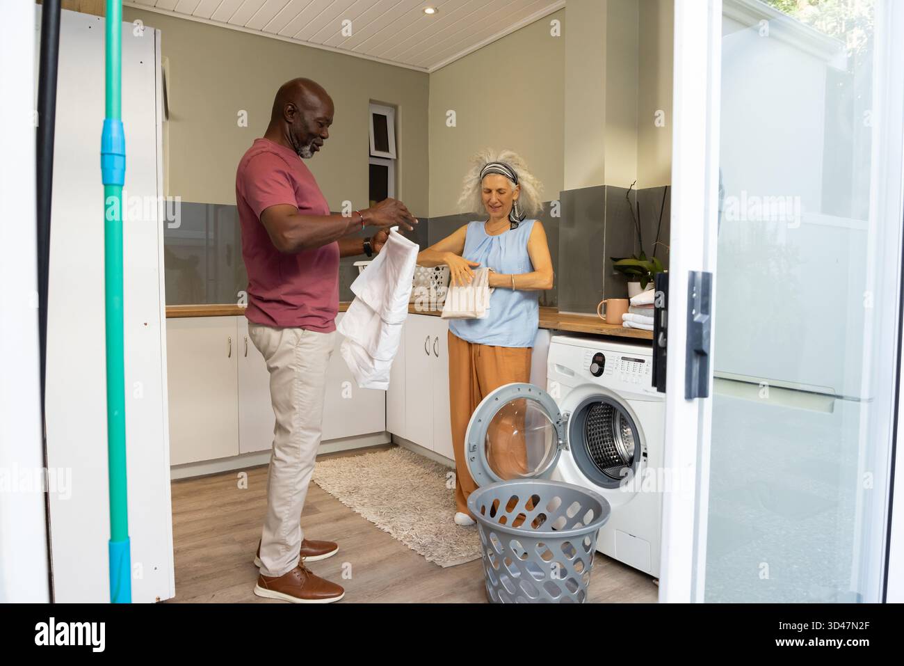 Lavanderia pieghevole di diverse coppie senior in una luminosa lavanderia casalinga, con cestino e asciugamani piegati Foto Stock
