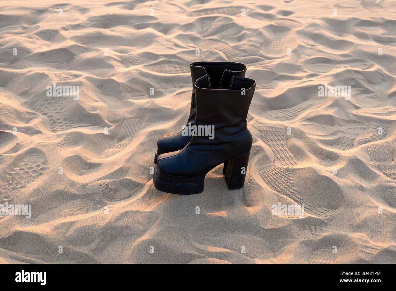 Doha, Qatar - 30 ottobre 2025: Un paio di eleganti stivali neri con tacco alto in piedi su morbida sabbia del deserto durante un sereno tramonto. Foto Stock