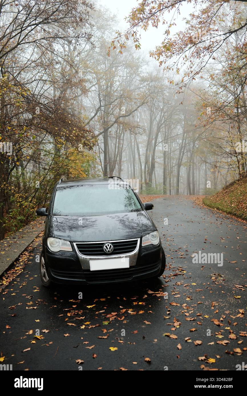 Auto Volkswagen Touran nera parcheggiata su una strada boschiva bagnata ricoperta di foglie autunnali con il tempo nebbioso. Un ambiente di viaggio tranquillo con una luce soffusa. Sopot, Serbia - Foto Stock