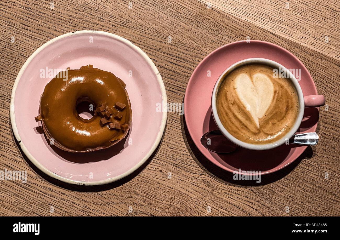 Ciambella ad anello ricoperta di cioccolato su un piatto rosa insieme a un caffè cappuccino, servito al Brammibal's Donuts di Berlino. Le ciambelle sono fatte a mano da Vegan - Immagine stock catturata con smartphone
