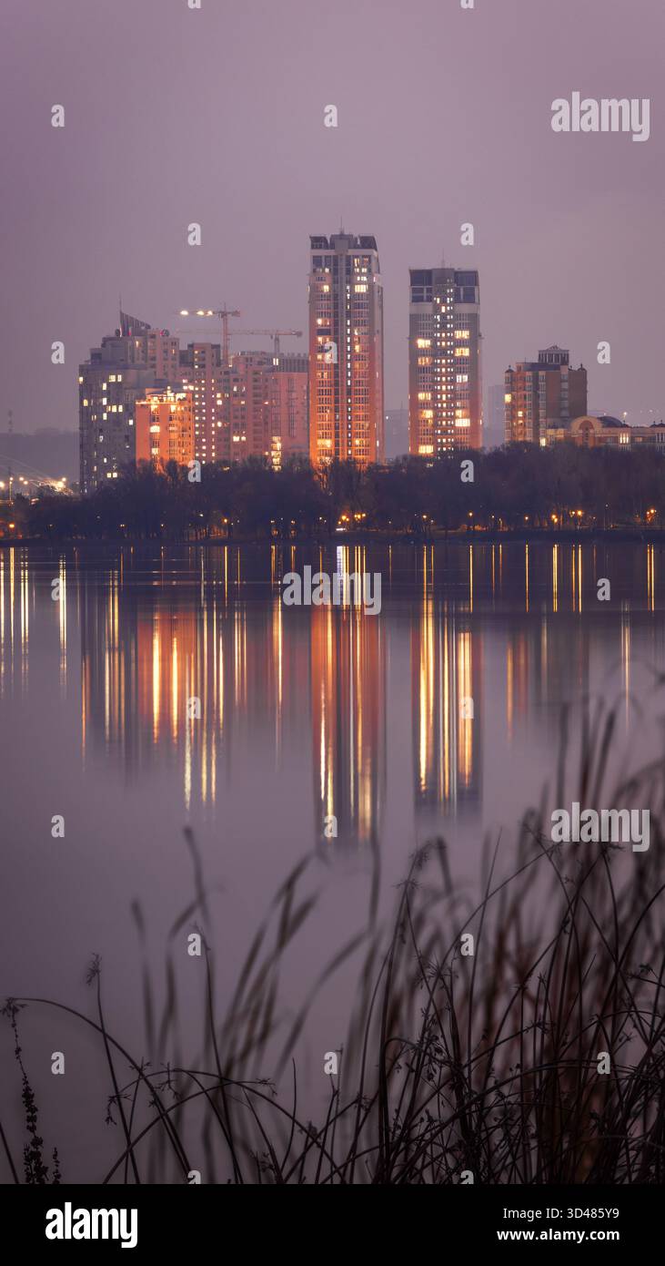 Vista serale dell'argine di Obolon a Kiev in una giornata nuvolosa. Le luci della città e gli alti edifici si riflettono nel tranquillo fiume Dnipro Foto Stock