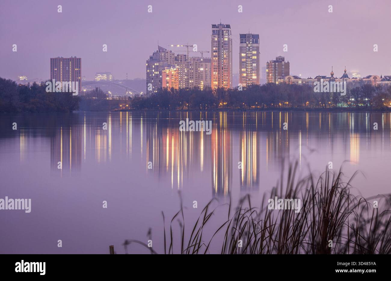 Vista serale dell'argine di Obolon a Kiev in una giornata nuvolosa. Le luci della città e gli alti edifici si riflettono nel tranquillo fiume Dnipro Foto Stock