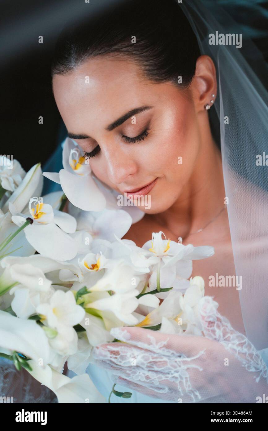 La sposa regge delicatamente un bouquet di orchidee bianche, gli occhi chiusi, abbracciando il momento con un'espressione serena. I suoi guanti in pizzo e il velo aggiungono un tocco di ele Foto Stock