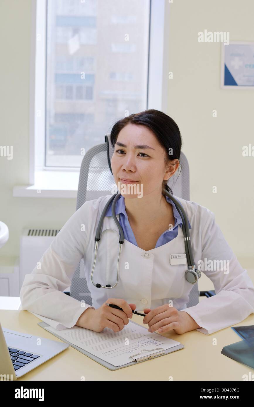 Donna asiatica di mezza età dottore seduto alla scrivania tenendo la penna, indossando stetoscopio, guardando leggermente a lato, lavorando con documenti medici in ufficio moderno con sfondo finestra Foto Stock