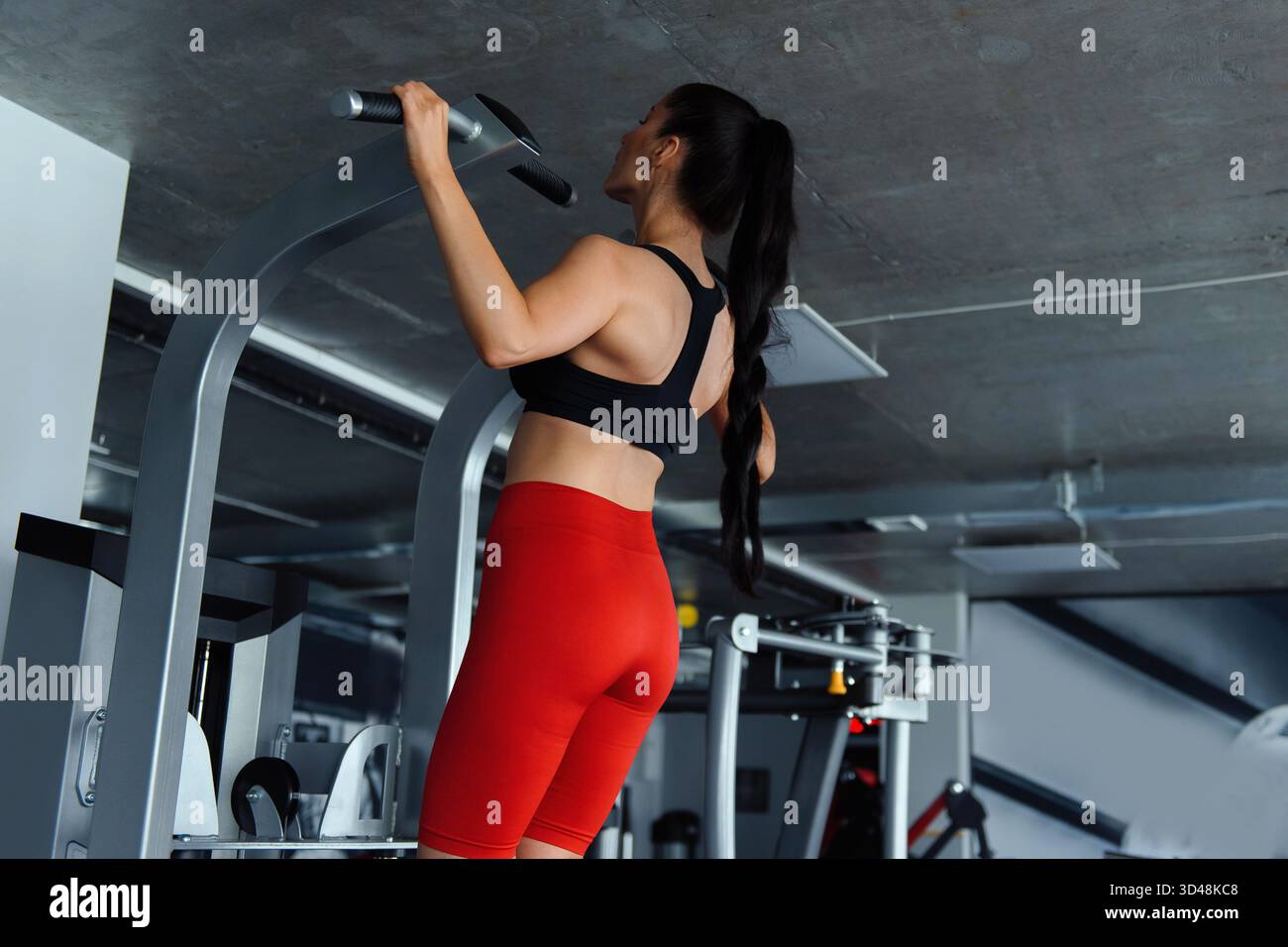 Vista posteriore di giovane donna con corpo muscolare in reggiseno nero e pantaloncini rossi braccia da allenamento su palestra. Una robusta brunetta che si esercita regolarmente per rimanere Foto Stock