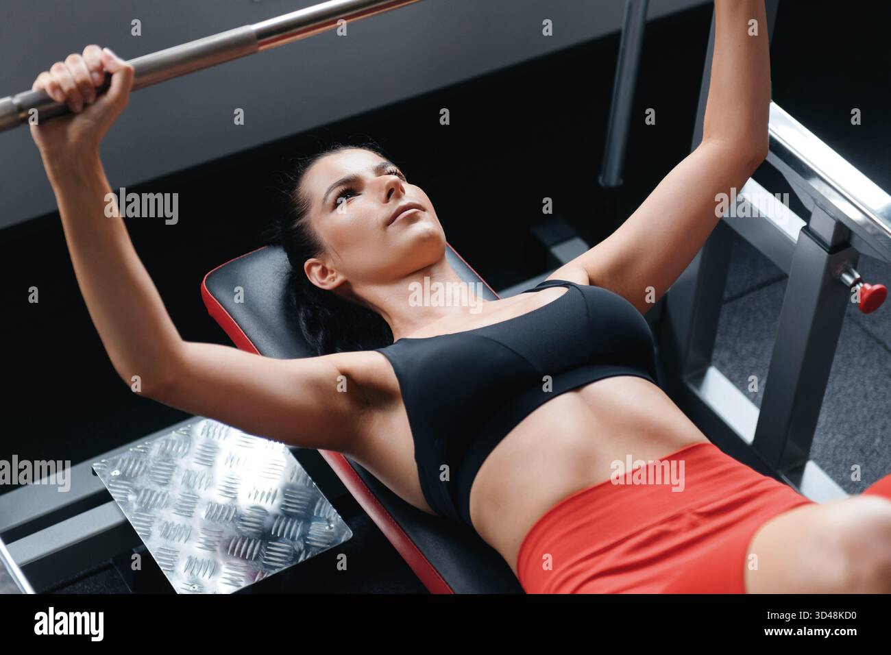Bella giovane donna in reggiseno nero e pantaloncini rossi sdraiati su panca e braccia da allenamento con barbell in palestra. Concept sport e allenamento. Foto Stock