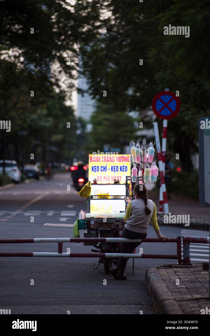 Un venditore ambulante vietnamita aspetta accanto al suo luminoso carrello di spuntini mobile al crepuscolo, catturando un momento tranquillo ma autentico della vita quotidiana urbana. Foto Stock