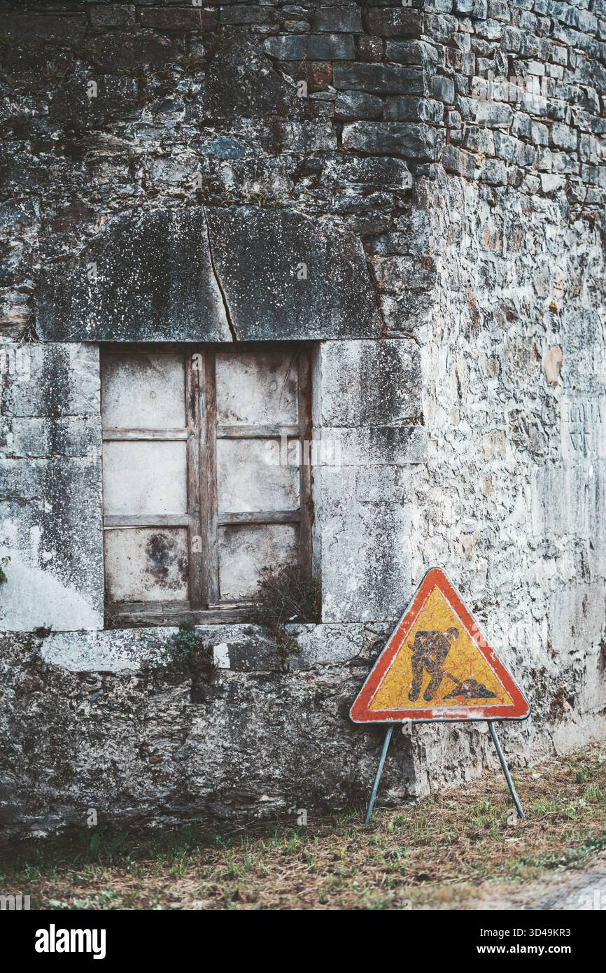 Cartello di costruzione intempestivo appoggiato su un vecchio muro di pietra con una finestra di legno sigillata. Texture rustica, toni attenuati e decadimento naturale creano un nos Foto Stock