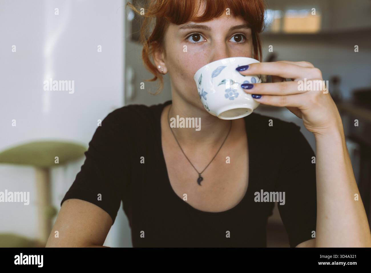 Ritratto giovane donna con i capelli rossi legati in panino seduto a tavola, tenendo la tazza e il piattino in mano, bevendo tè verde, infusione di erbe, caffè, mattina Foto Stock