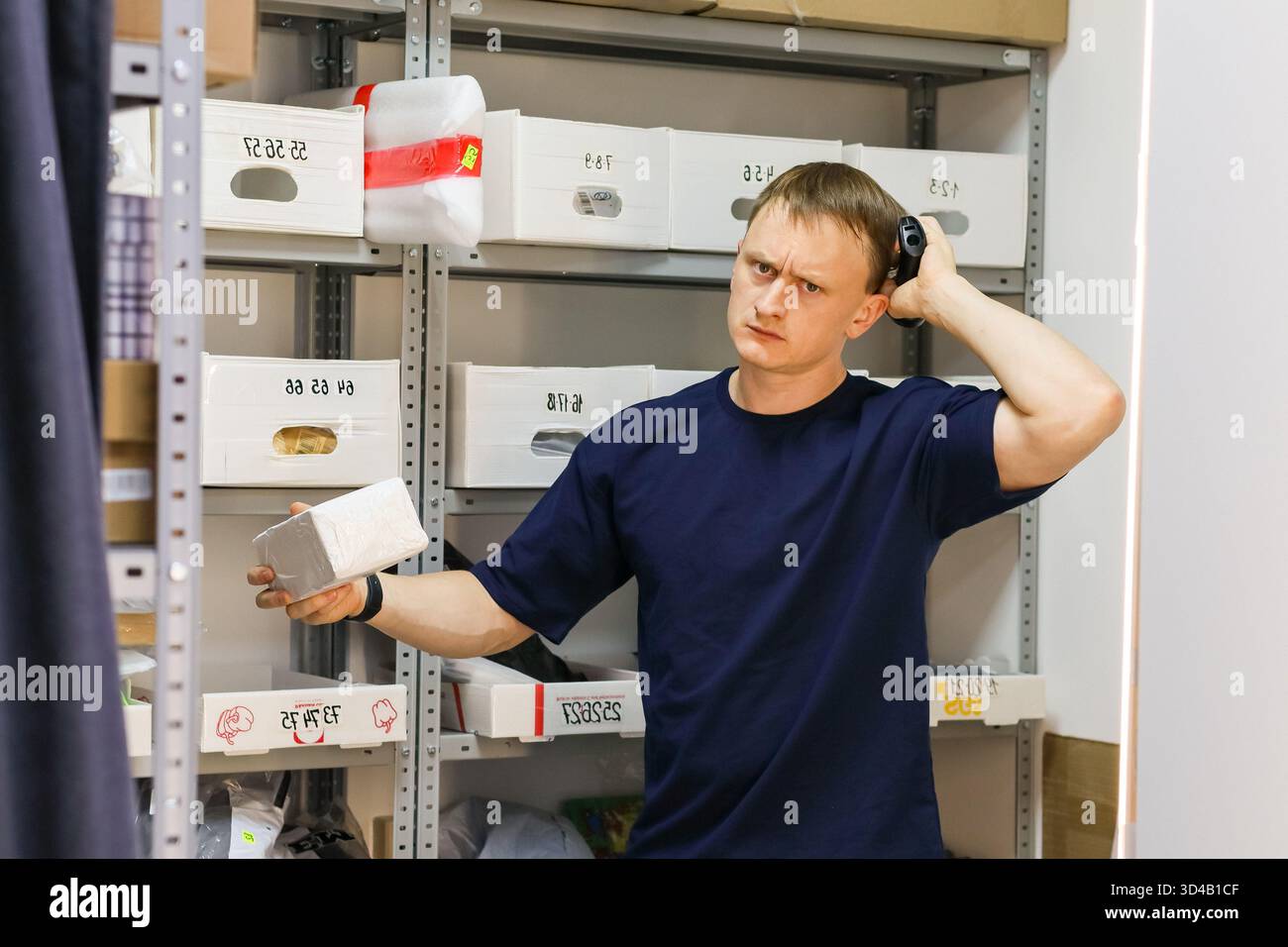 Dipendente che ha riflettuto con lo scanner nel magazzino. L'uomo in camicia scura si concentra profondamente afferrando lo scanner portatile accanto ai ripiani etichettati. Foto Stock