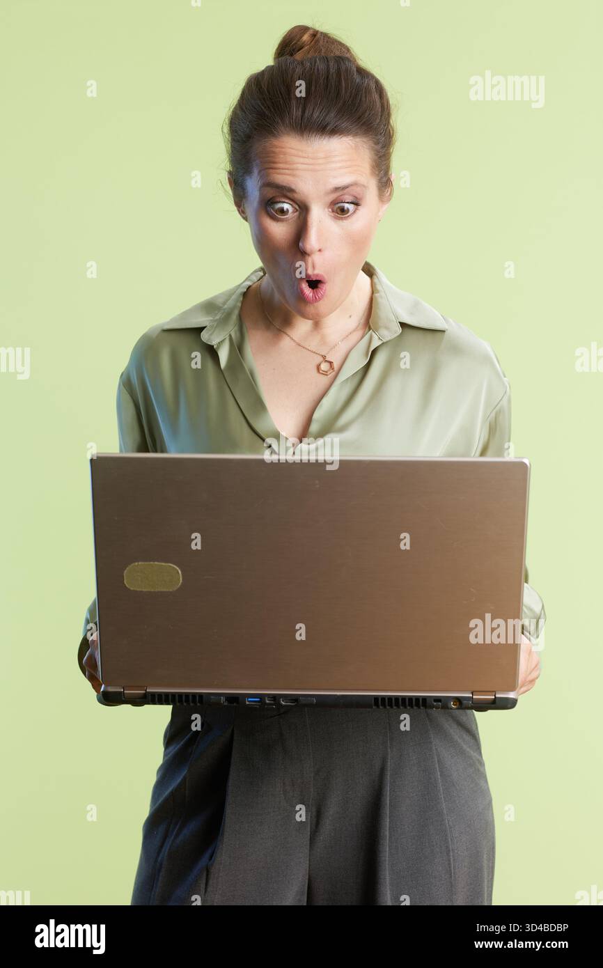 Una donna sorpreso guarda il proprio notebook in stato di shock o di eccitazione, indossando una camicetta verde e pantaloni grigi su uno sfondo verde chiaro. Foto Stock