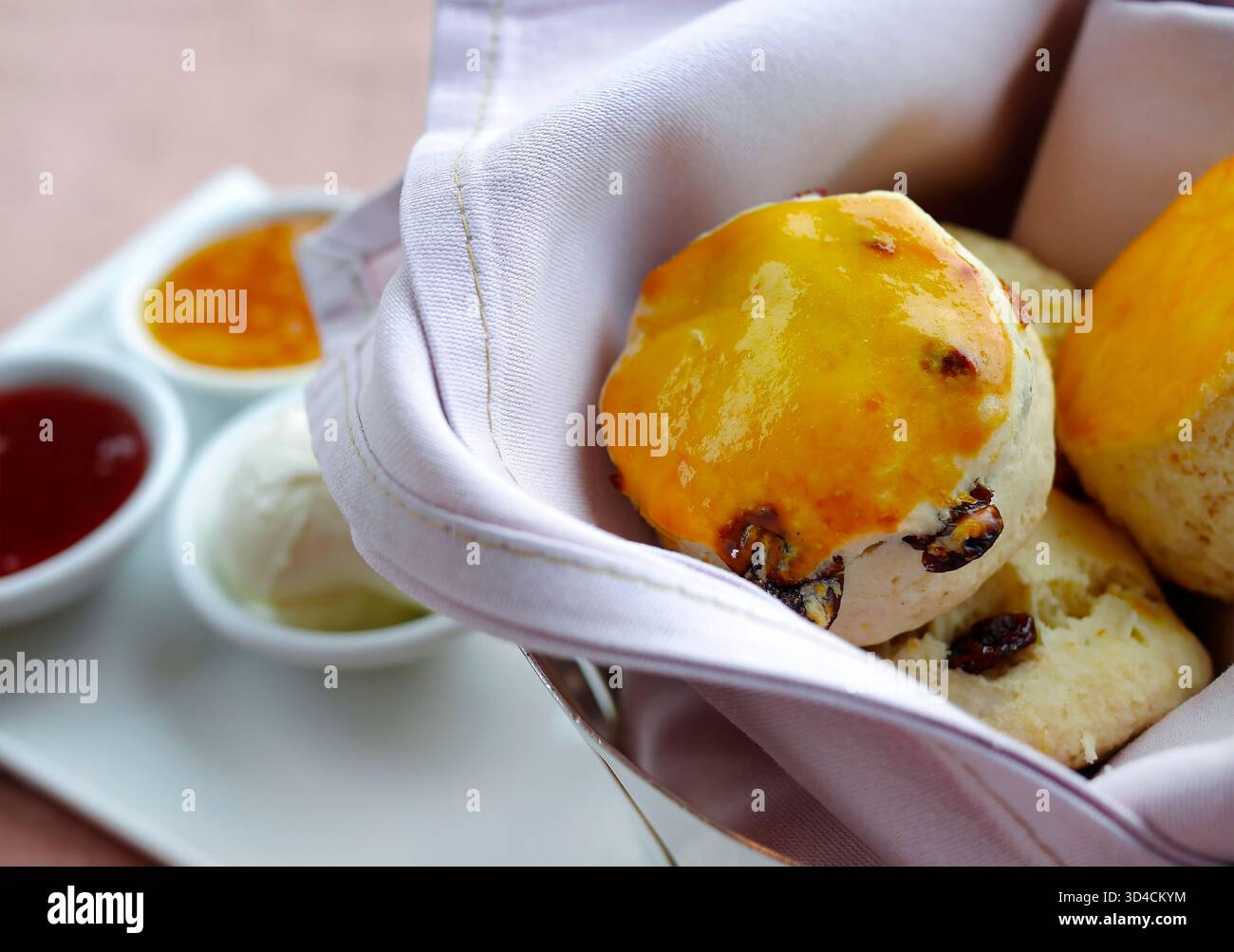 Primo piano di focaccine dorate appena sfornate servite in un cestino rivestito di tessuto, abbinate a marmellata di fragole, marmellata e panna. Pulito, luminoso e appe Foto Stock