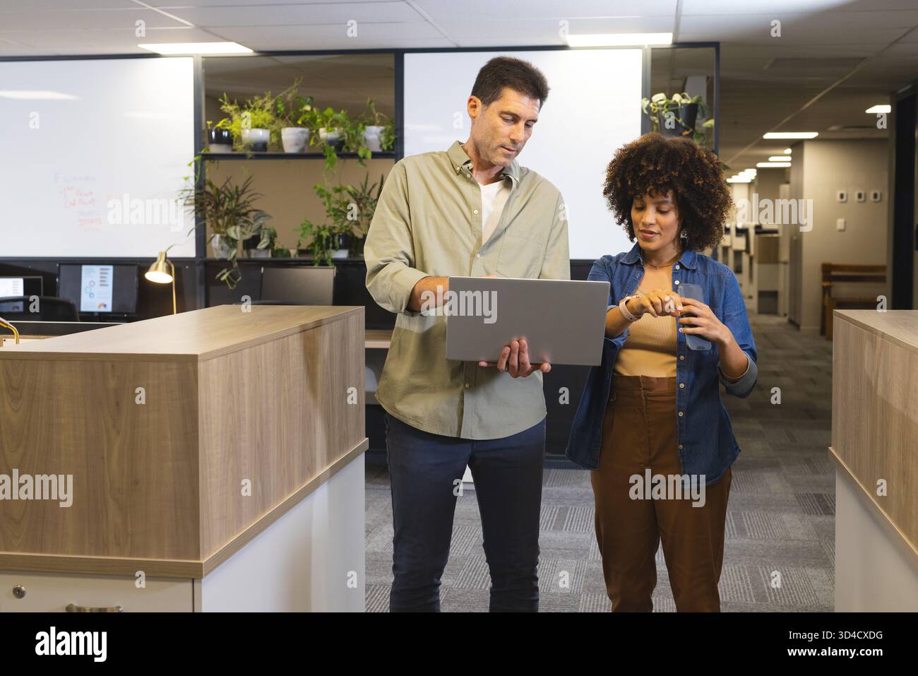 Colleghi diversi che esaminano i piani dei progetti nei corridoi degli uffici, con laptop e smartwatch Foto Stock