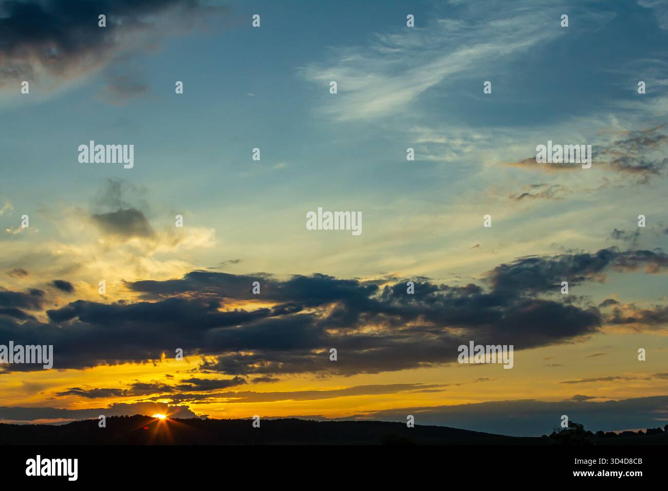 Uno splendido tramonto dipinge il cielo con sfumature di arancione e blu, mentre le nuvole si spostano pigramente sopra le colline ondulate creando una scena tranquilla nella campagna. Foto Stock