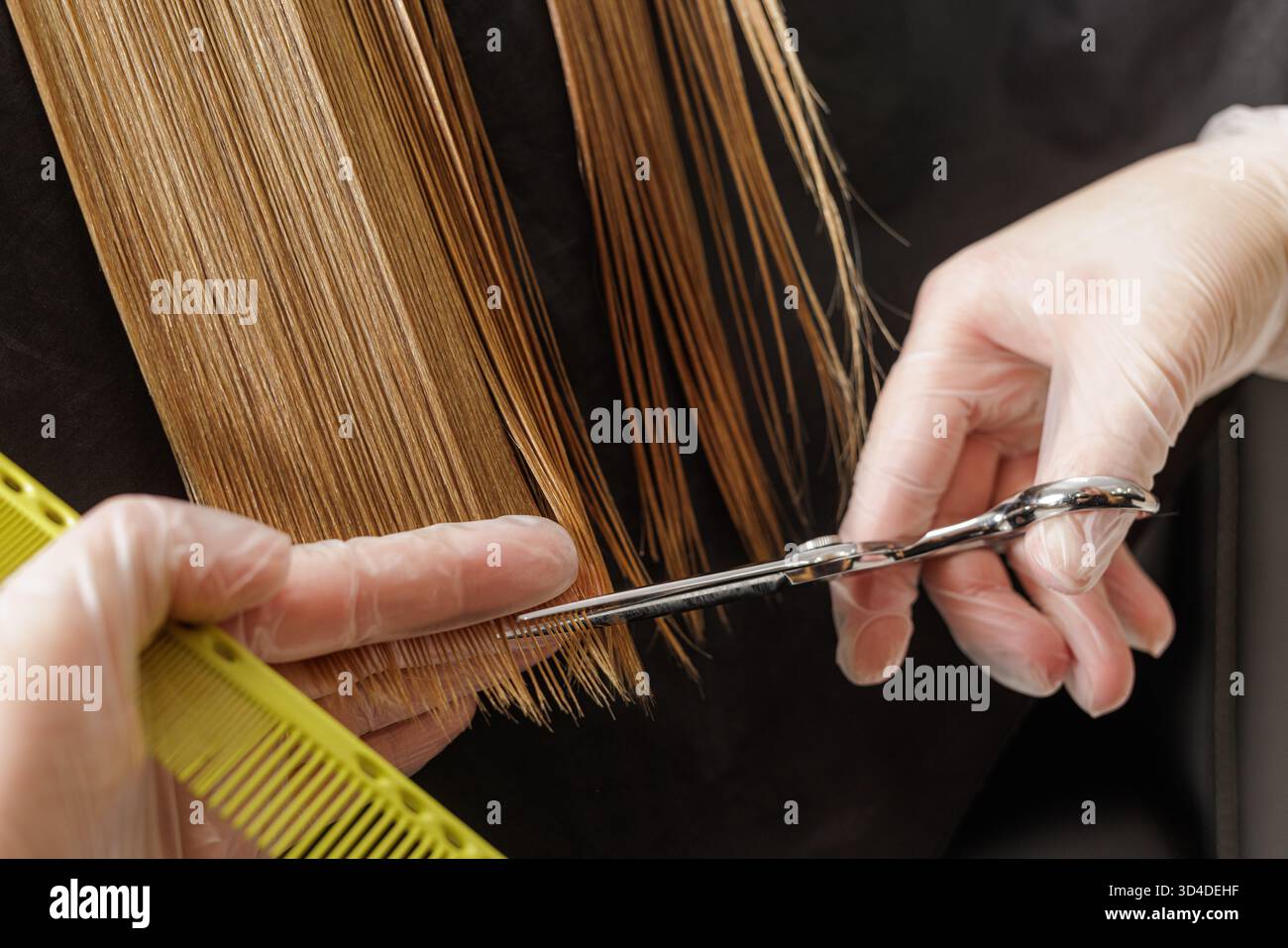 I guanti per parrucchieri rifiniscono i lunghi filamenti umidi con forbici e pettine, formando una linea retta precisa durante il servizio di un salone professionale. Foto Stock
