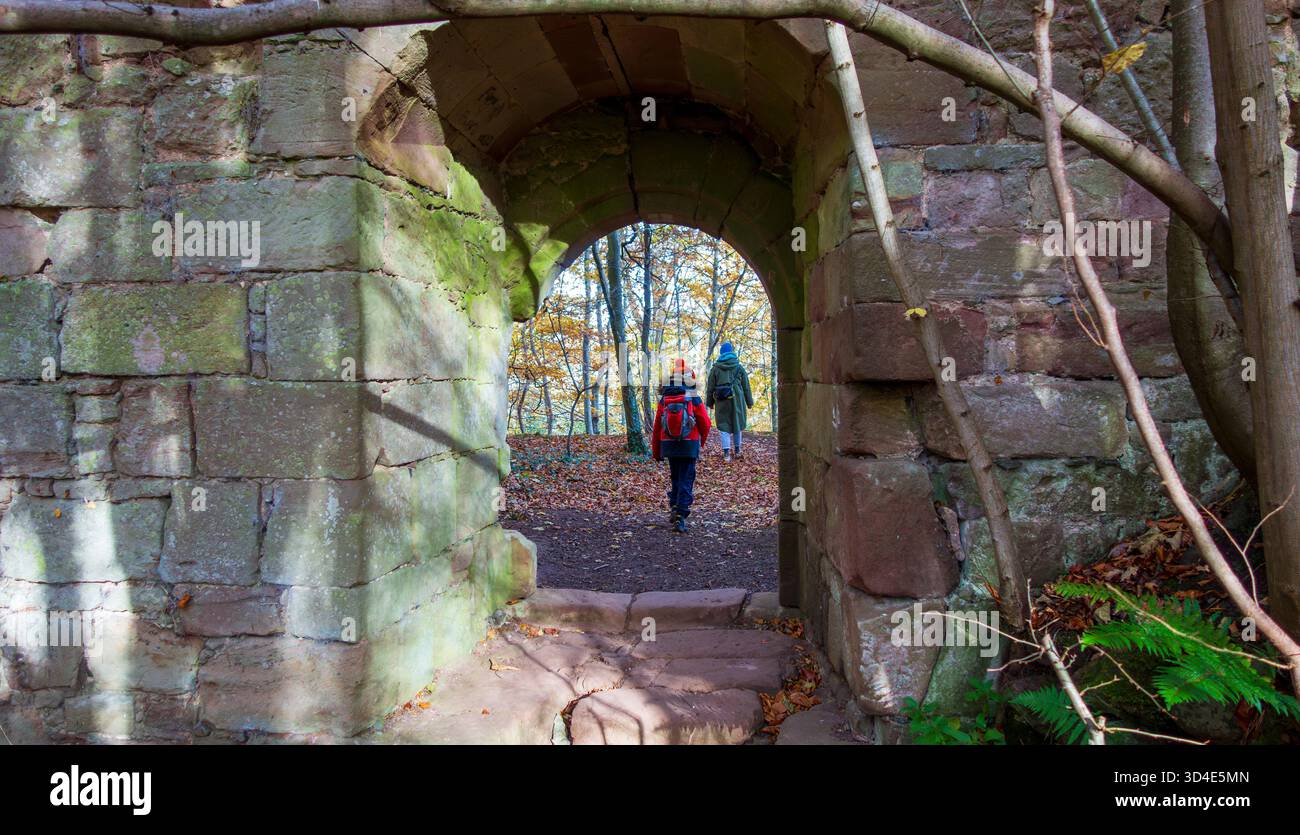 Vista dell'arco di pietra intemprata che incornicia un sentiero cosparso di foglie autunnali, che attira due figure in una scena boschiva dondolata dal sole, Haddington, Scotl Foto Stock