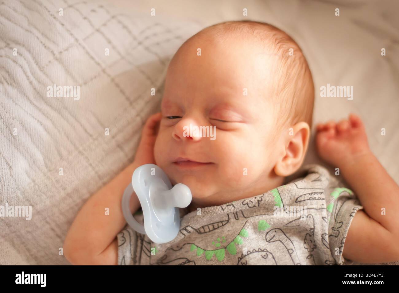 Un neonato dorme sonni su una coperta a motivi geometrici. Il bambino ha un succhietto vicino alla bocca e indossa una camicia grigia con i dinosauri stampati Foto Stock
