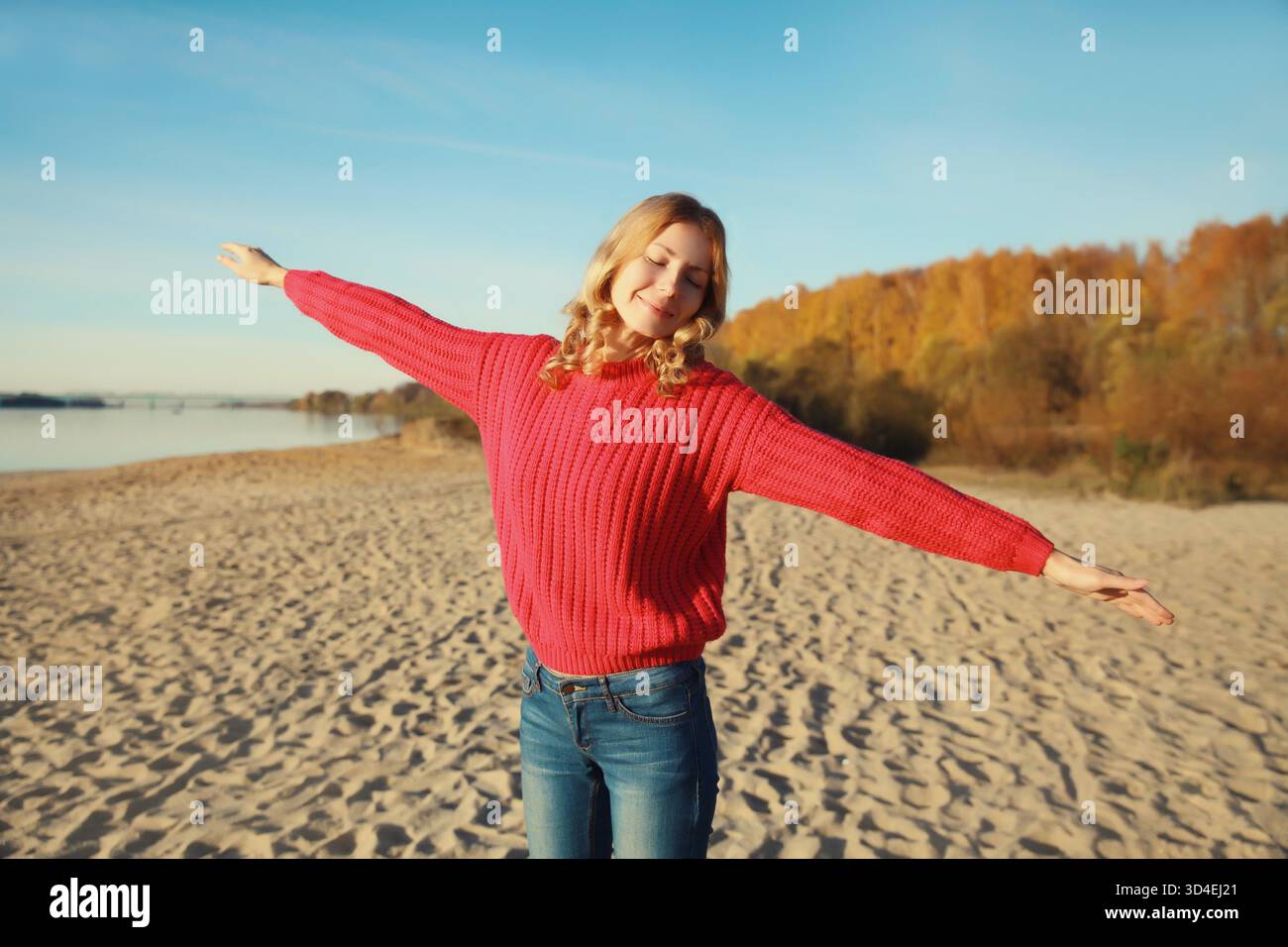 Giovane donna felice e rilassata che sorride camminando sulla spiaggia, godendosi il caldo sole, indossa un maglione in piedi sulla costa del mare, contro alberi con foglie gialle, sfondo naturale || modello rilasciato Foto Stock