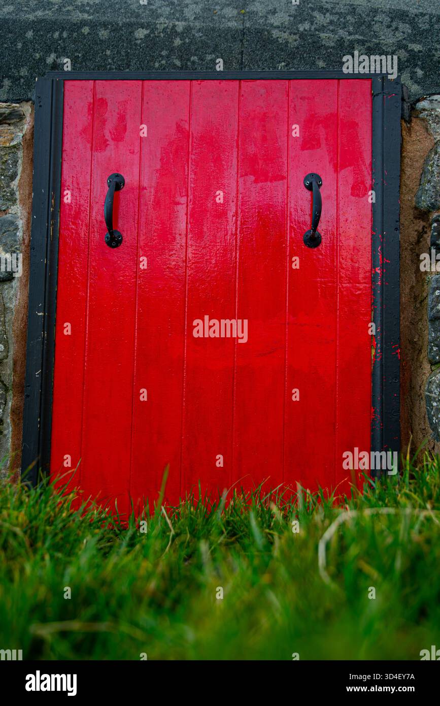 Una porta rossa di dimensioni medie con maniglie nere. erba in primo piano e la porta è su un muro di pietra Foto Stock