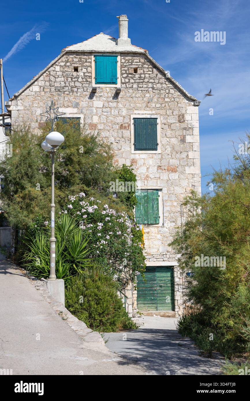 Una casa rustica in pietra con persiane blu turchese si trova accanto a un sentiero di ghiaia e lussureggiante vegetazione sotto un cielo blu cristallino. Perfetto per viaggi, architettura, Foto Stock
